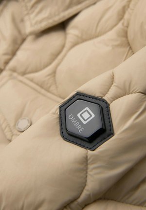 Tela de chaqueta acolchada beige con un parche hexagonal negro etiquetado "OMBRE" cosido en la manga.