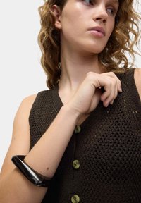 Junge Frau mit lockigem Haar, die ein schwarzes gestricktes ärmelloses Top und ein großes schwarzes Armband an ihrem linken Handgelenk trägt, vor einem weißen Hintergrund.