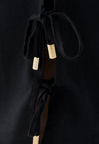 Prenda negra con una abertura lateral cerrada con lazos de tela negra y aplicadores metálicos de color dorado. Textura suave con un acabado liso.