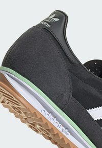 Zapatilla deportiva negra con parte superior de ante y malla, rayas blancas, acento en la entresuela en verde y suela de goma con un patrón de zigzag.