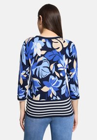 Femme aux longs cheveux foncés portant un haut bleu à motifs floraux avec des manches trois-quarts et un ourlet rayé, associé à un jean bleu.