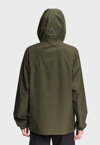Personne portant une veste à capuche vert olive vue de dos avec une lettre A grise imprimée sur la capuche.