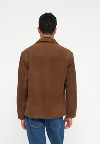 Brown corduroy jacket with a notched collar and long sleeves, featuring a clean back design and subtle texture. Paired with blue denim pants.