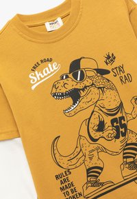 Keltainen puuvillateepaita, jossa on dinosauruksen grafiikka aurinkolaseilla ja lippiksellä, sisältäen tekstejä ja urheilullisen muotoilun.