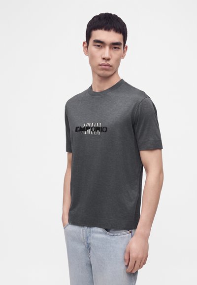 Jeune homme portant un t-shirt Armani Emporio gris foncé et un jean bleu clair, debout avec la main droite dans la poche devant un fond blanc.