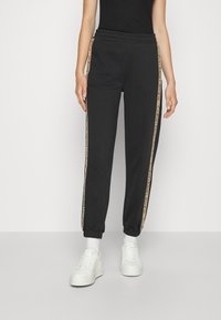 Černé tepláky mají elastický pas, zúžené lemy a boční pruhy s béžovým logem. Kombinovány s bílými teniskami a ponožkami nad kotník.