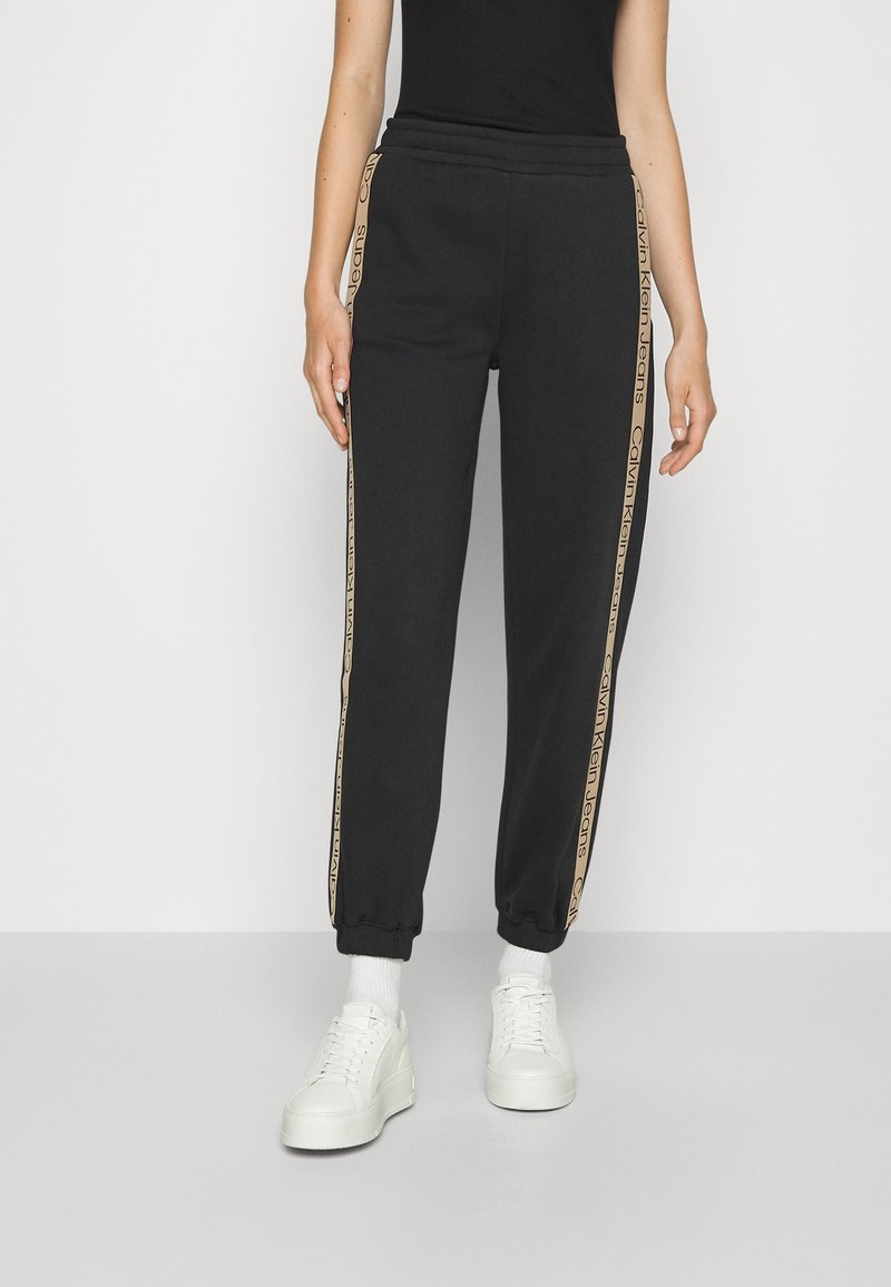 Černé tepláky mají elastický pas, zúžené lemy a boční pruhy s béžovým logem. Kombinovány s bílými teniskami a ponožkami nad kotník.