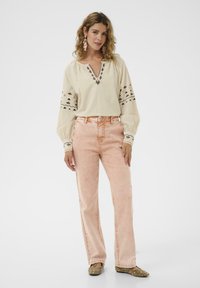 Kremkrāsas izšūta blūze ar V-veida kaklu un platajiem piedurknēm, kopā ar gaisīgi rozā izplestām džinsa biksēm. Modelis valkā dekoratīvas auskarus un plakanas kurpes.