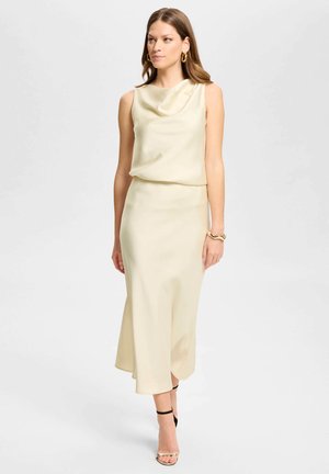 Donna con abito in raso senza maniche color crema con scollo drappeggiato, orecchini a cerchio dorati, braccialetto e sandali con tacco alto e cinturino nero, in piedi su sfondo bianco.
