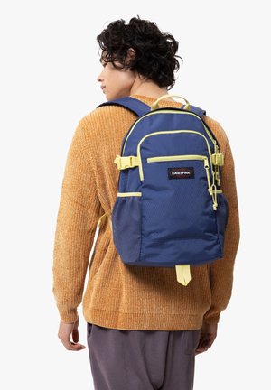 Navy blue Eastpak backpack with yellow accents, textured fabric, two front zip pockets, adjustable straps, and a top handle.