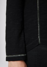 Zwart shirt met lange mouwen, uitgevoerd in een gestructureerde stof met contrasterende zilverkleurige stiksels langs de mouwzoom en zijnaden.