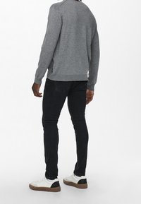 Homme portant une chemise à manches longues grise, un jean slim noir, des baskets blanches avec des talons noirs, debout de dos face à l'appareil photo sur fond blanc.