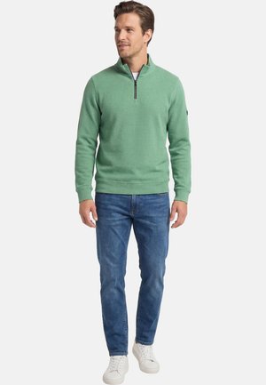 Uomo con maglione verde menta con cerniera a quarto di giro, jeans blu e scarpe da ginnastica bianche, in piedi e che guarda verso sinistra.