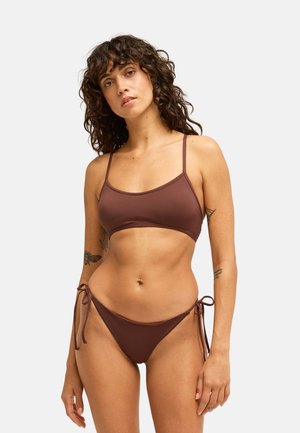 Femme aux cheveux bouclés et foncés portant un bikini marron avec des bretelles fines et des liens latéraux, debout devant un fond clair uni.
