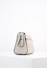 Piccola borsa a tracolla in pelle beige con bordi neri, cerniera argentata e una tasca laterale con zipper, posizionata su una superficie bianca contro uno sfondo bianco.