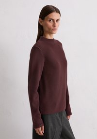 Brązowy sweter teksturowany z wysokim kołnierzem, długimi rękawami i luźnym fasonem, w połączeniu z szarymi spodniami. Miękka tkanina, minimalistyczny design.
