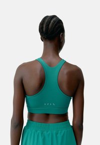Reggiseno sportivo verde con design a dorso scoperte, caratterizzato da una texture liscia e un branding sottile sul retro. Abbinato a pantaloni corti verdi coordinati.