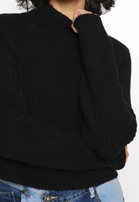 Czarny prążkowany sweter dziany z wysokim kołnierzem, długimi rękawami i luźnym krojem, charakteryzujący się fakturowaną powierzchnią oraz podwiniętymi mankietami, zestawiony z niebieskimi dżinsami.