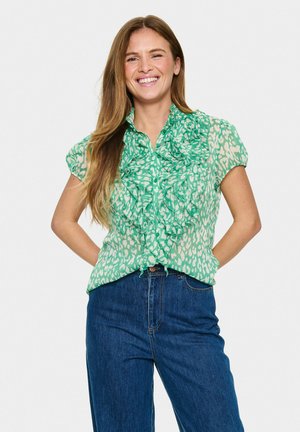 Green, patterned blouse with ruffled detail at collar and buttons, short sleeves, paired with dark blue denim jeans.