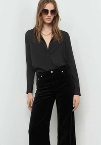 Camisa negra de botones combinada con pantalones de terciopelo negros de pierna ancha. La modelo lleva accesorios como gafas de sol de gran tamaño y un collar de cadena dorada.