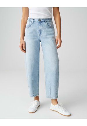 Persoon draagt lichtblauwe high-waisted rechte spijkerbroek, witte sneakers en een wit mouwloos topje, staand op een effen lichte achtergrond.