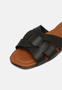 Sandale en cuir noir avec des brides tressées, bout carré et semelle intérieure en cuir marron. Présente des coutures contrastées et une semelle extérieure plate et texturée.