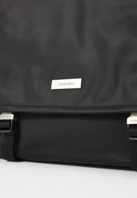 Sac à dos en tissu noir avec deux bretelles noires et une plaque en métal argenté avec le logo Calvin Klein centrée sur le rabat.
