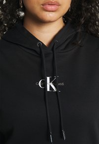 Sudadera negra con capucha y cordones, que presenta un logo blanco "CK" y el texto "CALVIN KLEIN JEANS" en la parte frontal. Textura de tela suave.