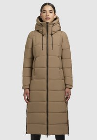 Vrouw in lange beige gewatteerde winterjas met capuchon, ritssluiting aan de voorkant en zijritsventilaties, staand tegen een effen lichte achtergrond.