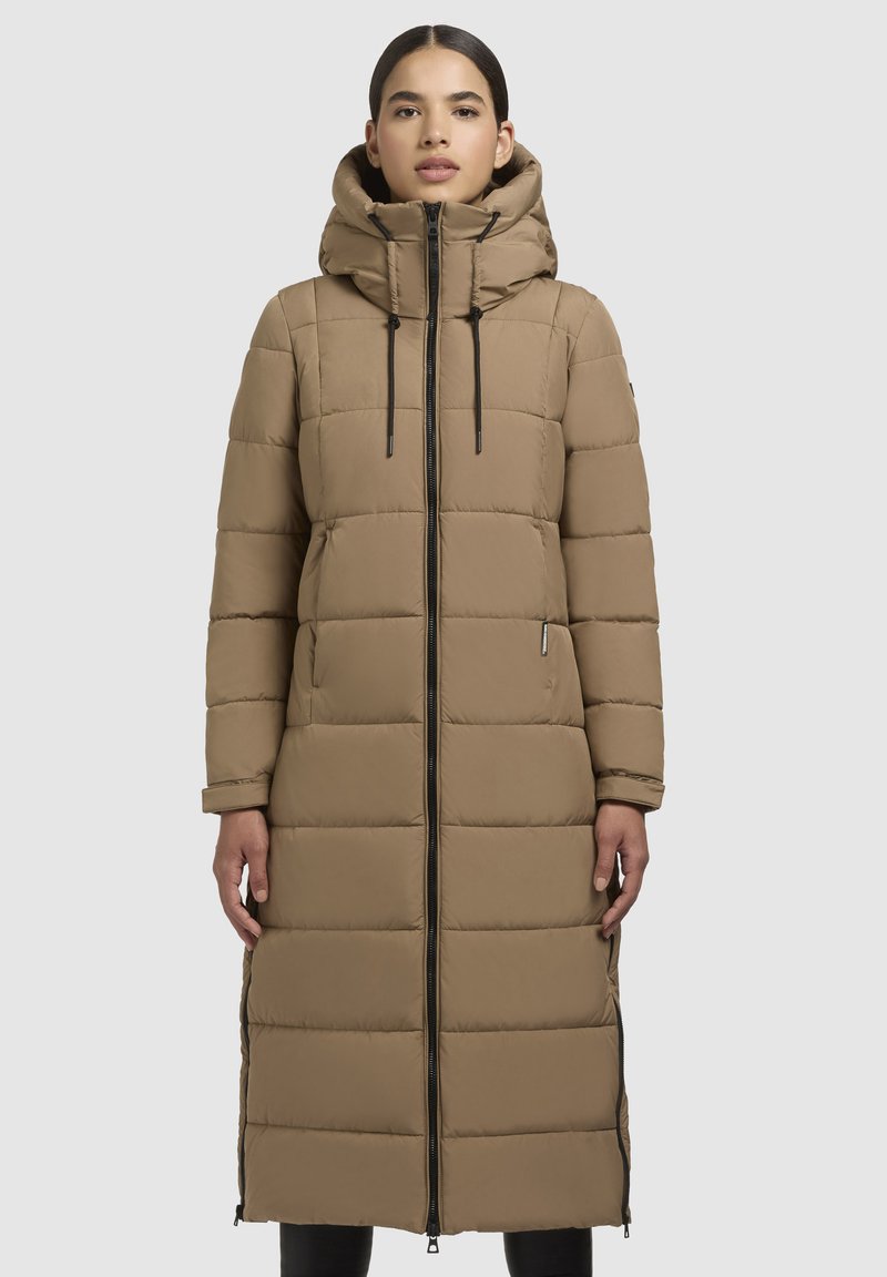 Vrouw in lange beige gewatteerde winterjas met capuchon, ritssluiting aan de voorkant en zijritsventilaties, staand tegen een effen lichte achtergrond.