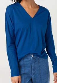 Kvinna i en långärmad blå V-ringad topp instoppad i högmidjade blå denimjeans mot vit bakgrund.