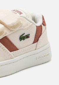 Sneaker in tessuto beige con dettagli marroni, cinturini in velcro e logo coccodrillo verde Lacoste. Parte superiore testurizzata con suola in gomma bianca.