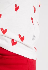 Biała koszulka z wzorem czerwonych serc, krótki rękaw i metka z boku. Zestawiona z czerwonymi spodniami, prezentuje codzienny strój.