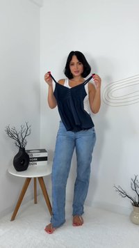 Námořnická modrá bezrukávová halenka s texturovaným předním dílem, knoflíky a splývavým lemem, kterou drží osoba v bílém tílku a světlých džínách.
