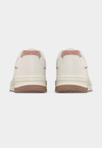 Λευκά αθλητικά παπούτσια με λεπτομέρειες σε ανοιχτό καφέ στη σόλα και τη φτέρνα, απεικονίζονται από πίσω σε απλό ανοιχτό γκρι φόντο.