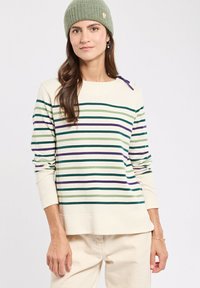 Femme portant un bonnet en tricot vert et un pull crème avec des rayures horizontales vertes et violettes, associé à un pantalon de couleur claire.