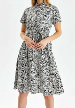 Kobieta ubrana w sukienkę o krótkich rękawach, z przodem na guziki, sięgającą połowy łydki, wykonaną z białej tkaniny w czarne, nieregularne plamy, z paskiem wiązanym w talii.