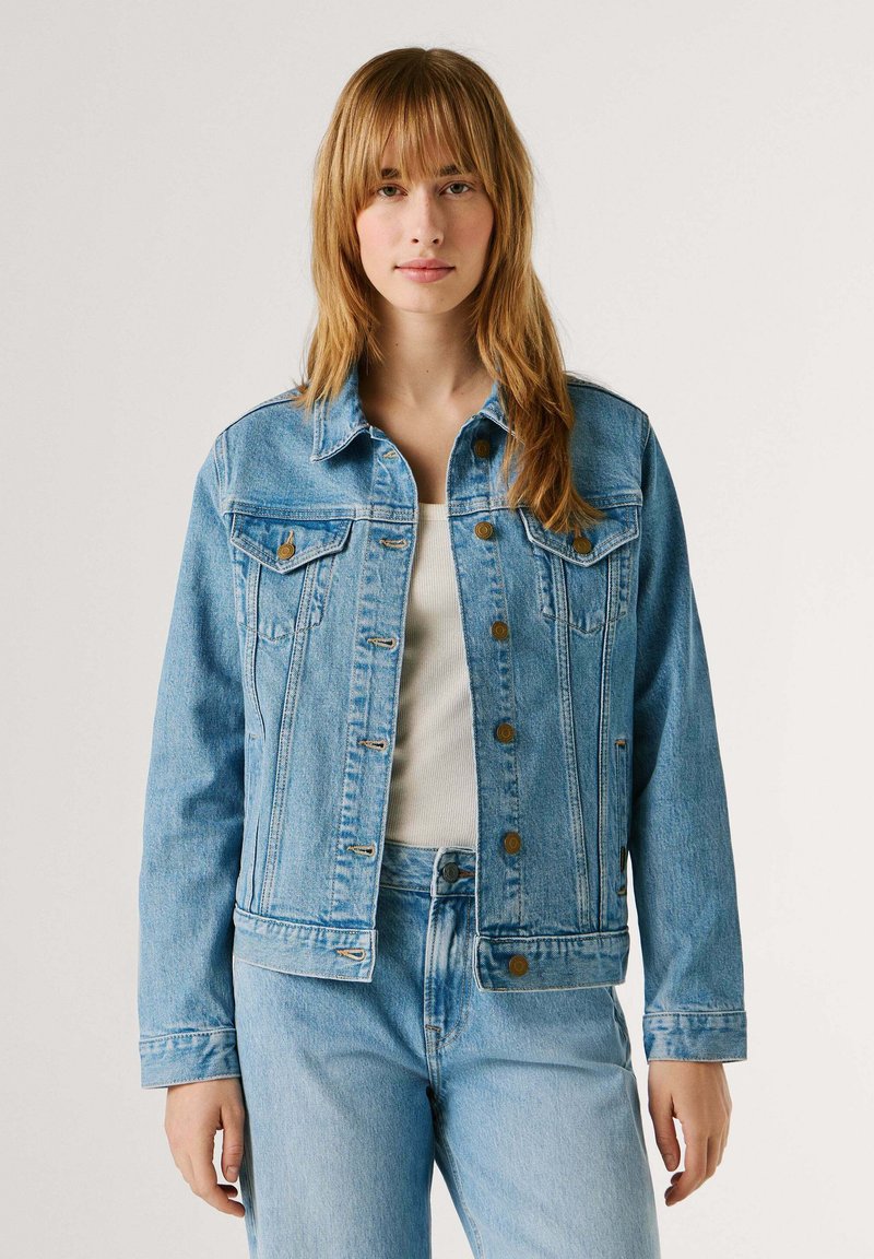 Ung kvinde med langt lyst brunt hår iført en lyseblå denimjakke og matchende jeans over en hvid top, stående mod en ensfarvet baggrund.