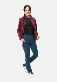 Sportoutfit bestehend aus einer bordeauxfarbenen Zip-up-Jacke, einem schwarzen, figurbetonten Top mit geometrischem Muster und tealfarbenen Stretchhosen mit seitlichen Taschen.