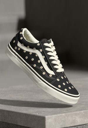 Zwarte lage sneaker met witte veters en zool, versierd met kleine witte en gele madeliefjes, zwevend boven een stenen oppervlak.