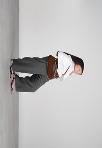 Femme avec de longs cheveux noirs, portant une blouse blanche, un pantalon large gris, des sandales noires et un sac à ceinture marron, se tenant de profil contre un fond uni.