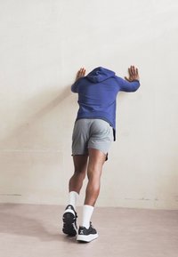Treningsklær: en blå hettegenser og grå shorts. Svarte sko med hvite detaljer på sålene, person vendt mot en lys vegg.