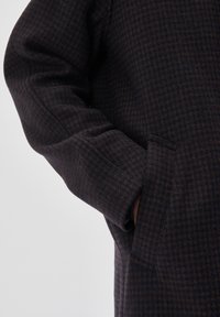 vlněný kabát v tmavom námorníckom odtieni s vzorom houndstooth. Má voľný strih, široké rukávy a bočné vrecká, pričom vyniká jemnou textúrou.