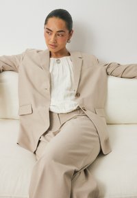 Ανοιχτό μπεζ κοστούμι με δομημένο σακάκι και παντελόνι σε άνετη γραμμή, συνδυασμένο με υφασματώδη λευκή μπλούζα και ασημένια κοσμήματα.
