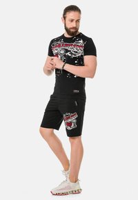 Schwarzes Grafik-T-Shirt mit roten und weißen Designs, kombiniert mit schwarzen Shorts mit ähnlichen Grafiken, getragen mit weißen Sportschuhen.