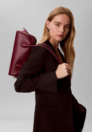 Mujer rubia con una chaqueta oscura lleva un bolso de cuero burdeos colgado del hombro sobre un fondo gris liso.