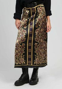 Jupe en satin noir avec des motifs paisley dorés et bordeaux, agrémentée de rayures géométriques. Portée avec des bottes de combat noires.