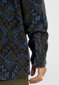Fleece-takki mustana, vihreänä ja sinisenä, jossa on geometrisia kuvioita. Täydennetty napitusyksityiskohdilla hihassa ja tekstuurilla, pehmeä pinta.