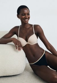 Beiger gepolsterter BH mit dünnen Trägern, kombiniert mit schwarzen Bikini-Shorts mit einem mehrfarbigen Bund; glatte Stofftextur.
