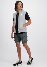 Graue ärmellose Zip-Weste über einem schwarzen T-Shirt, kombiniert mit dunkelgrauen Cargo-Shorts und schwarzen Sneakers mit weißen Akzenten und Taschen.
